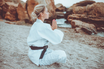 Attractive female martial artist in white kimono with black belt sits on sand and meditates on a wild beach