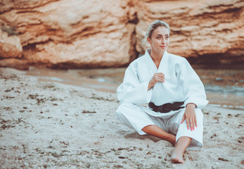 Attractive female martial artist in white kimono with black belt sits on sand wild beach
