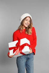 Happy young woman in Santa hat and sweater with gift box on light grey background. Christmas celebration