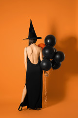 Young woman wearing witch costume with balloons for Halloween party on yellow background