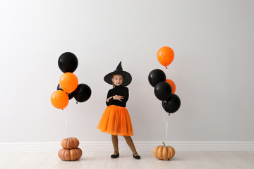 Cute little girl with balloons and pumpkins wearing Halloween costume near light wall