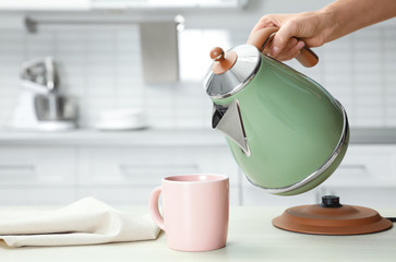 Woman pouring water from modern electric kettle into cup at wooden table in kitchen, closeup