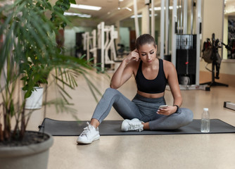 Young sport woman in sportswear using smartphone while sitting on mat in the gym. Workout break. Healthy lifestyle concept