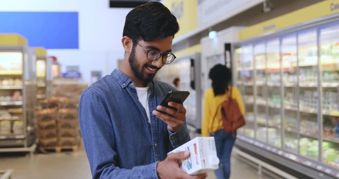 Young Arabian smiled guy in glasses taking a photo of milk or translating a text on it in the supermarket during shopping.