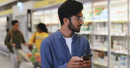 Arabian young cheerful and good looking guy texting and chatting on the smartphone in the supermarket while buying food.