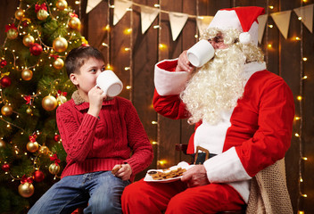 Santa Claus and child boy drinking tea, eating cookies, talking and sitting indoor near decorated xmas tree with lights - Merry Christmas and Happy Holidays!