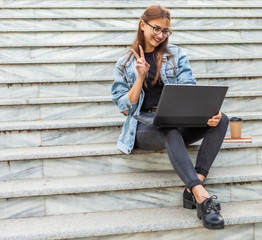 Online call. Young hipster woman in denim jacket communicates with friends on online conference with laptop while sitting on the stairs in the city