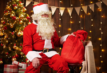 Santa Claus posing with bag of gifts, sitting indoor near decorated xmas tree with lights - Merry Christmas and Happy Holidays!