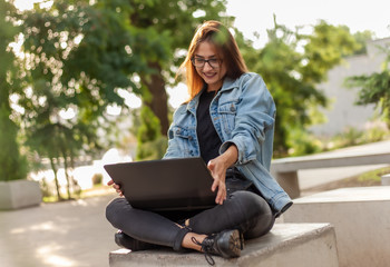 Young happy woman student in a denim jacket sitting at park and looks at the laptop screen. Distance learning. Online call. Modern youth concept.