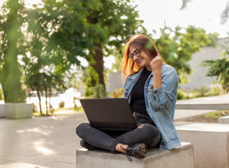 Young happy woman student in a denim jacket sitting at park and looks at the laptop screen. Distance learning. Online call. Modern youth concept.