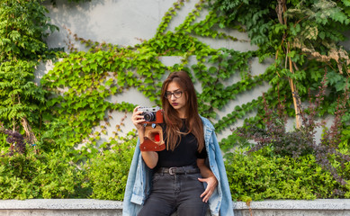 Young hipster woman in denim jacket and glasses enjoys film retro camera outdoors