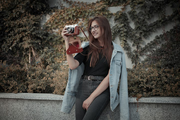 Young hipster woman in denim jacket and glasses enjoys film retro camera outdoors