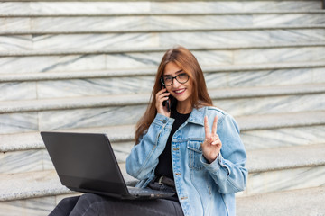 All in business. Young stylish woman in a denim jacket and glasses uses a laptop and talking on the phone while sitting on the stairs in the city. Remote work.