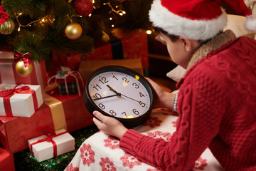 Teen boy waiting for Santa and watching the clock, lying indoor near decorated xmas tree with lights, dressed as Santa helper - Merry Christmas and Happy Holidays!