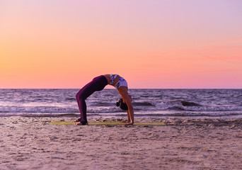 Adorable girl in sportswear is doing yoga by the sea. She bends, standing in the trough. Asana yoga pose. Beautiful portrait of yoga. Outdoors lifestyle portrait. Vacation, summer, relaxation.