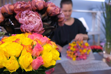 Floral business concept. Blur background with professional florist woman packaging roses boquet in flower shop. White and pink roses and peonies on table in vases. Working in floristic studio store.