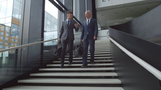 Wide shot of two Caucasian businessmen wearing elegant suits with bags in their hands going downstairs in airport while communicating
