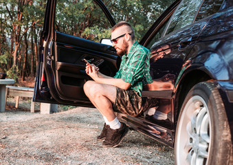 Cute bearded man with sunglasses sitting at car door looking at his phone