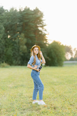 Fototapeta premium a girl in denim clothes and a hat stands in a field at sunset and holds a retro camera in her hands and smiles