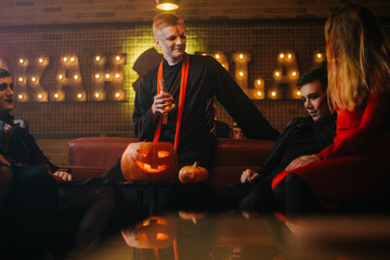Fototapeta premium Friends in costumes conversation at the halloween party. Beautiful girl in company of two guys in a cafe. Holiday carved pumpkin on table