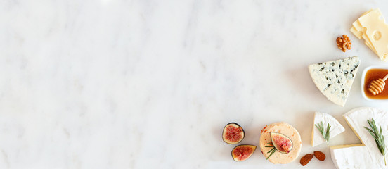 Corner border of a selection of cheeses, figs, nuts and honey. Above view banner on a white marble background with copy space.
