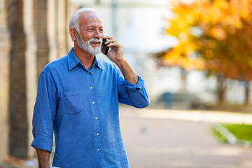 Happy bearded senior man talking by phone. Cheerful senior businessman talking by smartphone. Portrait of modern senior man speaking by phone outdoors