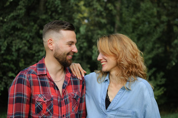 Obraz premium Girlfriend wearing blue casual jeans blouse is looking at handsome hipster boyfriend in red shirt walking in the park on green trees background