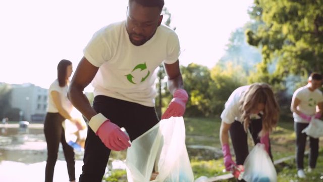 Focused afro-american volunteer in eco T-shirt searching picking up garbage into bags supporting nature movement with eco-friendly young team. Anti-pollution and ecology.