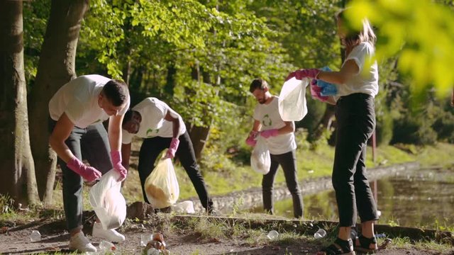 Team of hardworking young people collecting plastic trash in the forest at lake. Eco volunteers in white recycling T-shirts take part in cleaning operation. Teamwork. People and nature.