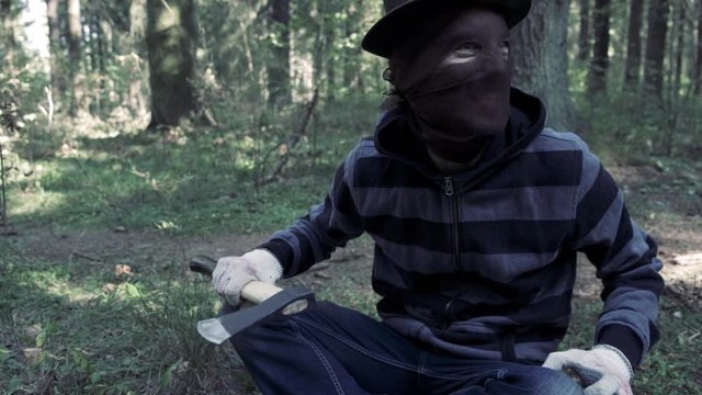 Serial maniac with axe in forest, bloody killer, murderer, mad man in scary mask and hat outdoor,  ready to kill. Halloween costume, horror concept.