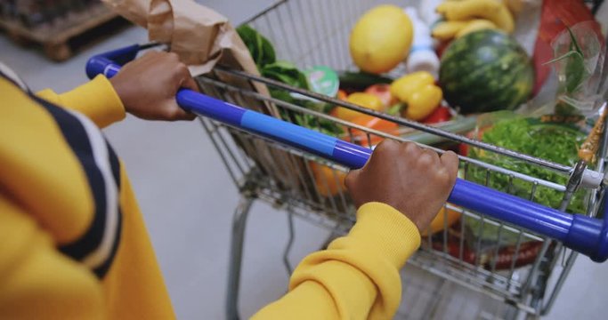 View from the top, from eyes of the shopper on the carriage with food, vegetables and fruit and African American female hands pushing it.