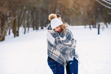 Fototapeta na wymiar Beautiful couple hugging each other and covered with checkered plaid while walking in snowy winter park.