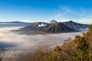 Plakat Mount Bromo volcano, island of East Java, Indonesia. Clouds cover the valley floor; Luhur Poten Temple at the foot of the cone. Gas from another volcano in the background rising against the blue sky. 