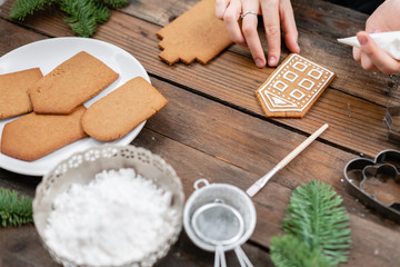 Icing of Christmas bakery. Woman decorating honey gingerbread cookies on wooden brown table. closeup, copy space. Blank biscuit gingerbread house, ready to decorate.