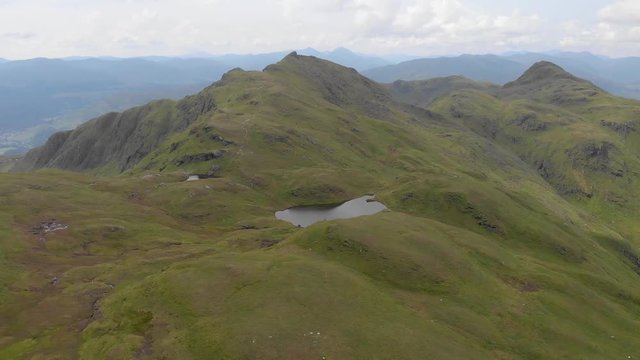 An aerial sideway right to left footage of a grassy mountain plateau rocky summit with small pond and mountain range in the background under a cloudy sky