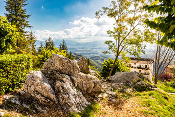 Fototapeta na wymiar Bellegra, Rome, Lazio, Italy - The panorama seen from Bellegra. The cloudy blue sky. Mountains and sunbeams. Glimpse of a public garden, with trees and spikes of rock.