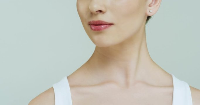 Close up of the bottom part of the face of young Caucasian woman with a smiling mouth and neck with shoulders on the white wall.