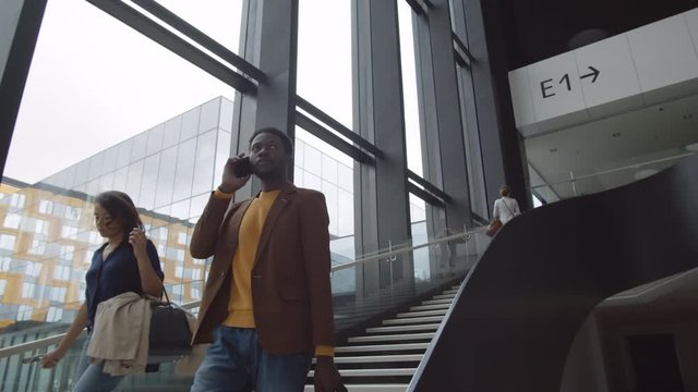 Medium shot of young African man wearing casual clothes with bag in his hand going downstairs in airport while talking on cellphone