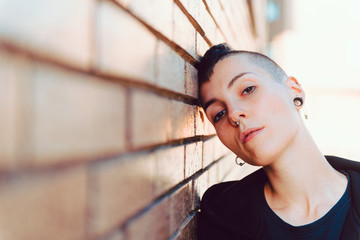 Young and attractive girl with shaved head and piercings. He has his head resting on a brown brick...