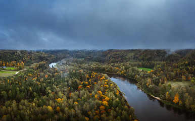 Valley of colorful trees covered fog at early mourning. Rainy weather with storm clouds in the sky. Picturesque panorama with river Gauja curving through the valley. 