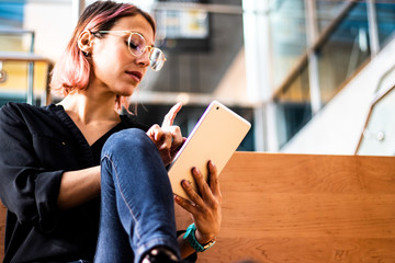 Smiling and pretty business woman using her digital tablet computer to read the news. She wears eyeglasses and blue jeans, casual style day. Technology app.