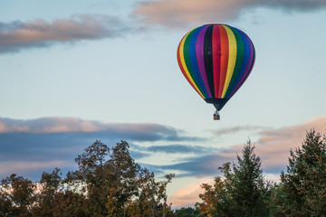 Rainbow hot-air balloon floats over grassy field and trees at sunrise