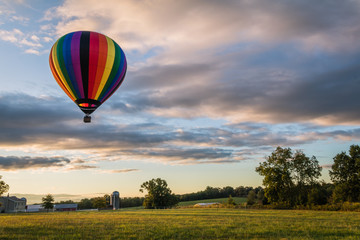 Rainbow hot-air balloon floats over farm field on a late summer morning as the sun rises