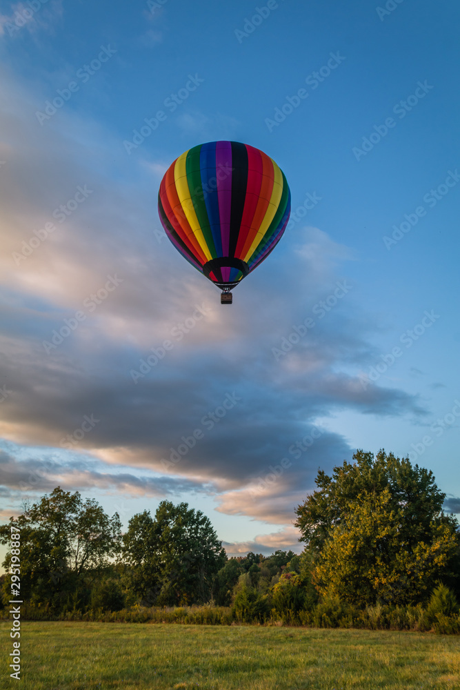 Poster Rainbow hot-air balloon floats over grassy field and trees at sunrise