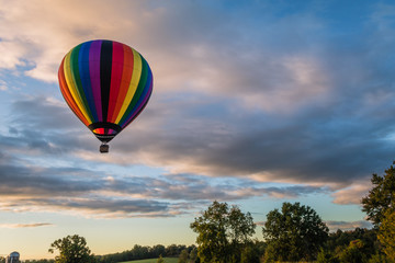 Rainbow hot-air balloon floats over grassy field and trees at sunrise
