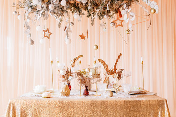 New Year mood, Christmas tree. Rectangular table covered with a gold tablecloth, decorated with candles with flowers for celebration. Festive still life by candlelight.