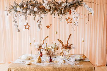 New Year mood, Christmas tree. Rectangular table covered with a gold tablecloth, decorated with candles with flowers for celebration. Festive still life by candlelight.