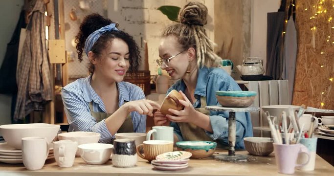 Two Caucasian young girls potters having fun, laughing cheerfully while examining ceramic cups and pods in the pottery studio.