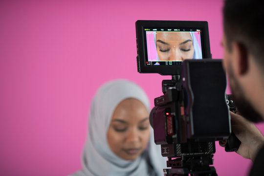 videographer in pink studio recording video on professional camera by shooting female muslim woman