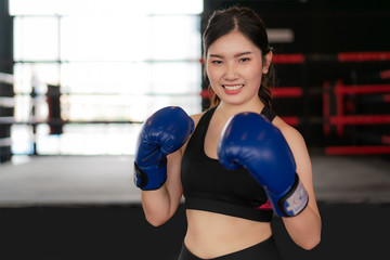 Fototapeta premium Young Asian Boxing fitness woman smiling happy wearing blue boxing gloves with Boxing Stadium in background. Portrait of sporty fit for healthy lifestyle Asian model of boxing gym concept..